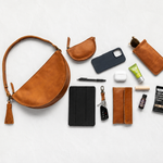 A curated flat-lay of brown leather accessories and small everyday items arranged on a light gray background, including a crescent shoulder bag with tassel, pouches, phone case, sunglasses holder, wallet, key fob, pen, earbuds case, and small toiletry items.
