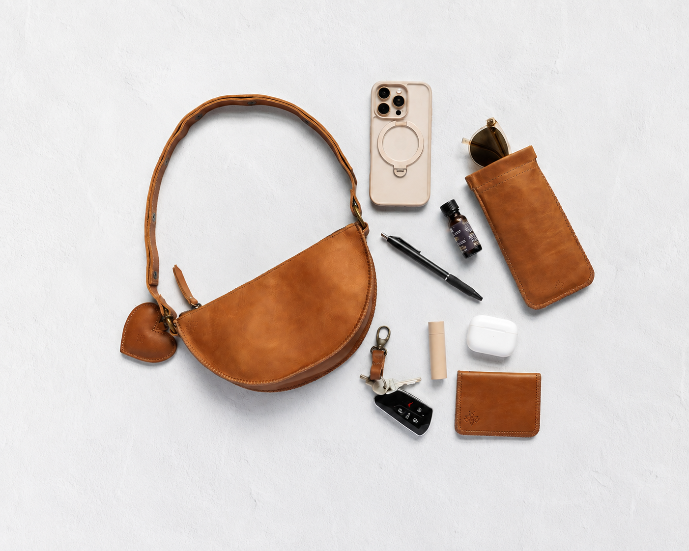 A warm tan leather half-moon shoulder bag with a matching leather key fob and card holder, arranged in a styled flat-lay with various accessories on a light gray textured background.