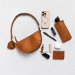 A warm tan leather half-moon shoulder bag with a matching leather key fob and card holder, arranged in a styled flat-lay with various accessories on a light gray textured background.