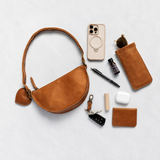 A warm tan leather half-moon shoulder bag with a matching leather key fob and card holder, arranged in a styled flat-lay with various accessories on a light gray textured background.