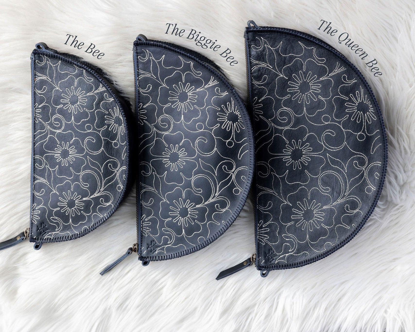 Three floral-patterned pouches labeled 'The Bee', 'The Biggie Bee', and 'The Queen Bee' on a white fluffy surface.