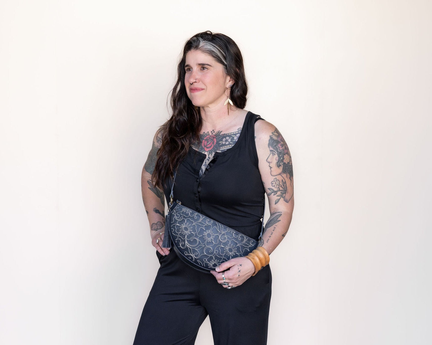Woman wearing a black sleeveless top and dark pants with a patterned belt against a plain background
