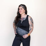Woman wearing a black sleeveless top and dark pants with a patterned belt against a plain background