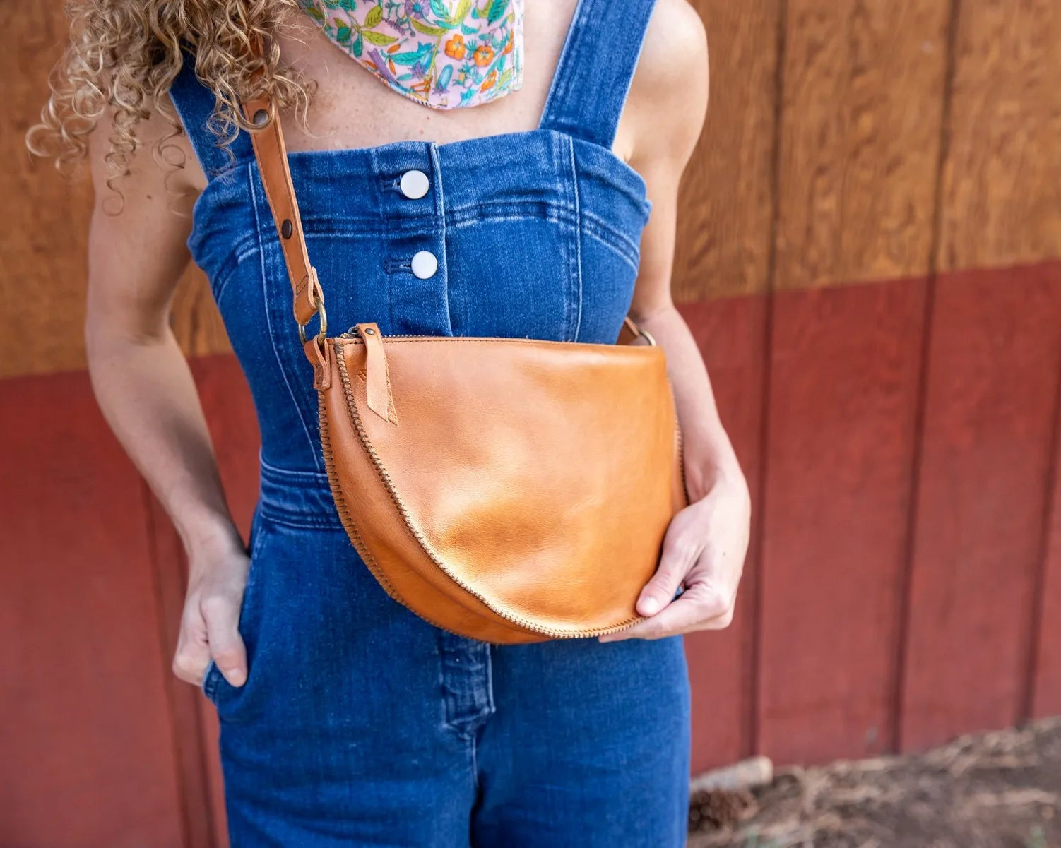 Person wearing blue overalls holding a brown leather bag against a wooden wall.