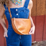 Person wearing blue overalls holding a brown leather bag against a wooden wall.