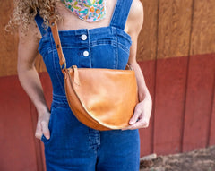Person wearing blue overalls holding a brown leather bag against a wooden wall.